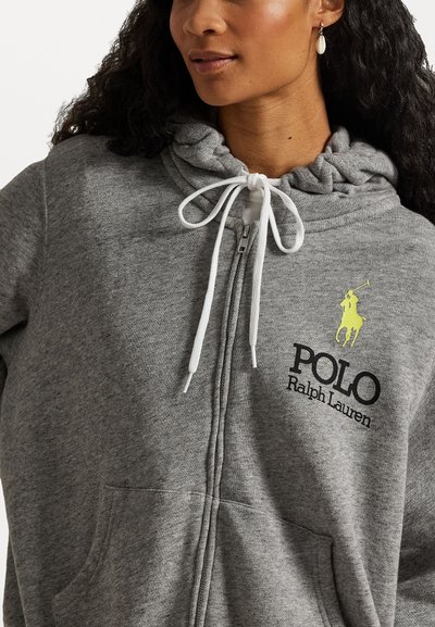 Pelēks zip-up džemperis ar auklu un dzeltenu polo logotipu, kā arī melnu tekstu. Gluda auduma un priekšējās kabatas.