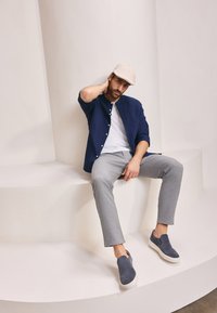 Marine knoop-overhemd, witte t-shirt, lichtgrijze broek en marine instappers. Model zittend op een gebogen wit platform met een neutrale achtergrond.