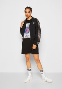 Black cropped jacket with patterned sleeves, graphic t-shirt, and black skirt. White socks with black stripes and chunky white sneakers.