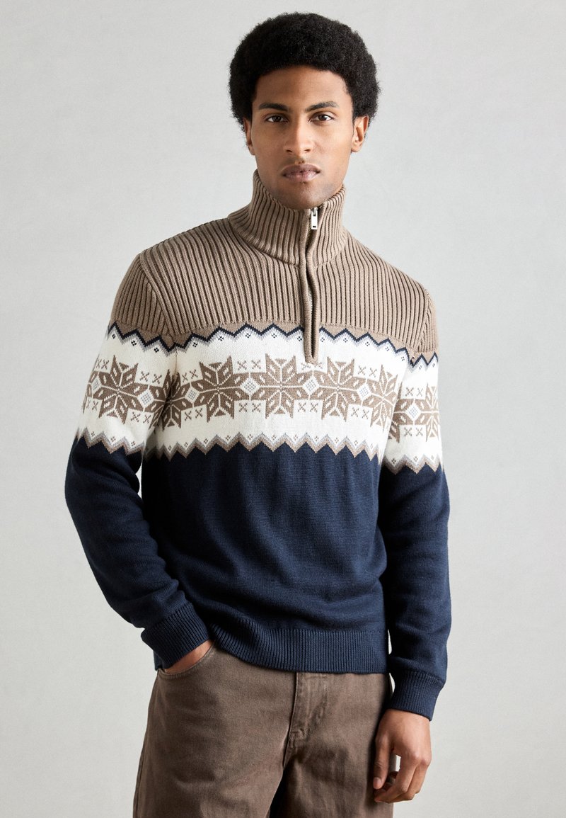 Camisola de malha com gola alta, apresentando um padrão de floco de neve em bege e branco sobre um corpo azul-marinho. Textura canelada na gola e nos punhos.