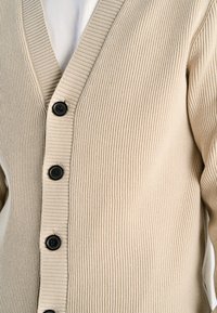 Beige geribbelde cardigan met een V-hals, vijf zwarte knopen aan de voorkant en een gestructureerde stof. Ontworpen voor een aansluitend silhouet.
