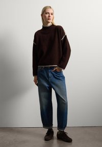 Une jeune femme se tient avec une main dans sa poche, portant un pull marron avec des bordures claires, un jean bleu ample retroussé aux chevilles et des baskets sombres.