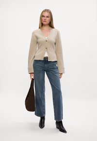 Beige cardigan med to knapper, brede mansjetter, kombinert med blå høyt liv jeans og svarte hælboots til ankelen. Brun veske i hånden.
