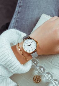 Hopeakuituinen rannekello, jossa on valkoinen kellotaulu, kultaiset yksityiskohdat ja mustat tuntimerkit, yhdistetty kultaketjuun neulepuseron peittämällä ranteella.