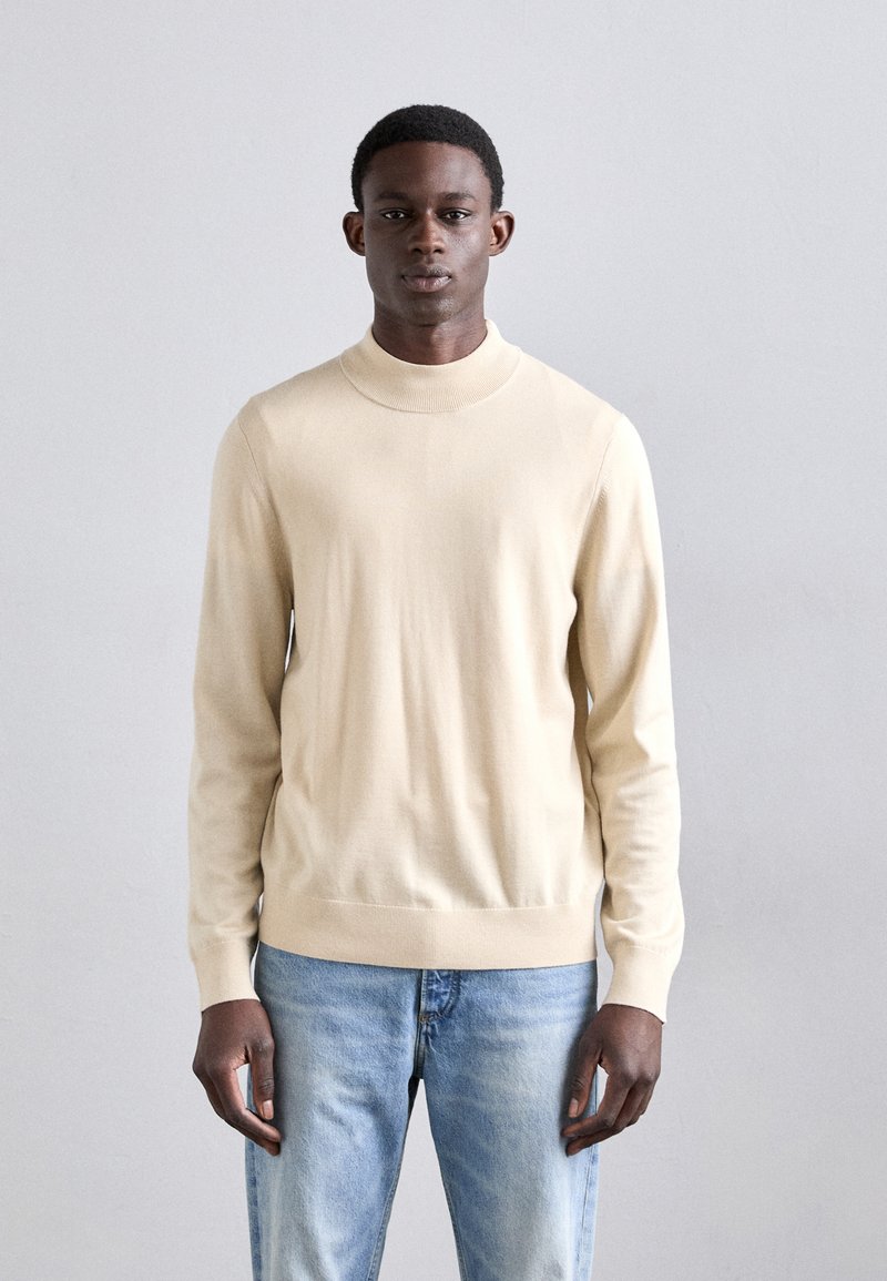 Beige gestrickter Pullover mit hohem Kragen, langen Ärmeln, gerippten Bündchen und einem taillierten Saum. Getragen mit hellblauen Jeans.