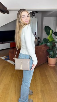 Gaiši pelēka crossbody soma ar strukturētu virsmu, zelta ķēdes siksna un pušķa detaļa. Nēsāta mājas interjera fona priekšā.