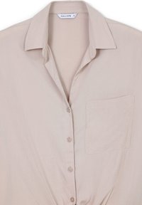 Blusa colletto a maniche corte in tessuto beige chiaro, con tasca frontale e chiusura a bottoni. Texture liscia con un leggero lucido.