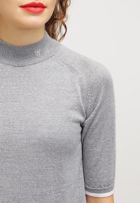 Woman wearing a gray short-sleeve knit top with a high collar embroidered with a small "W" on the left side.