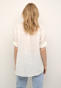 Hvid linnedbluse med oprullede ærmer og en afrundet kant. Har et rygstykke og et let, struktureret stof.