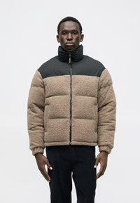 Geacă puffer cu un corp din fleece texturat, de culoare maro, și o secțiune superioară din nailon negru. Prezintă un guler înalt și un fermoar complet în față.