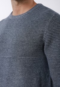 Homme portant un pull en maille bleue texturée avec un motif côtelé sur la partie haute de la poitrine et les manches et une maille lisse sur la partie basse de la poitrine.