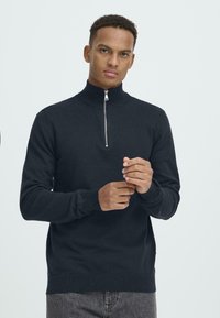 Crna zip-up dukserica s visokim ovratnikom. Mekani pleteni materijal, prilagođen rukavi, rebrasti manžeti i rub. Minimalistički dizajn, bez uzoraka ili ukrasa.