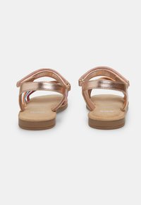 Roségoldene metallische Sandalen mit flacher Sohle, verstellbaren Riemen und weicher Innenauskleidung. Die Rückansicht zeigt ein offenes Design und minimale Verzierungen.