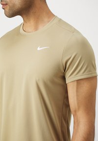 Beige treningstrøye med kort erm, laget av strukturert stoff, har en hvit Nike-logo på brystet.