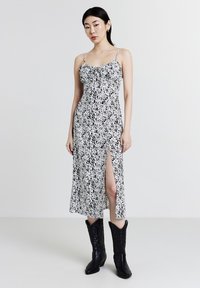 Donna che indossa un vestito midi a pois bianco e nero con uno spacco elevato e stivali da cowboy neri, in piedi contro uno sfondo bianco semplice.