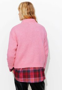 Pull en tricot rose avec un col montant, présentant une coupe ample et des poignets côtelés. Porté par-dessus une chemise à carreaux multicolore à couches.