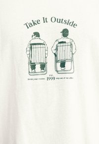 T-shirt blanc avec un graphique vert représentant deux personnages assis sur des fauteuils de jardin. Le texte indique « Take It Outside » et « Break your routine. est. 1999. »