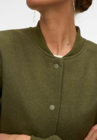 Femme portant une veste en laine vert olive boutonnée avec un col côtelé et une boucle d'oreille pendante, les bras croisés.