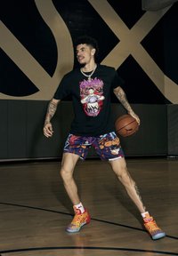 Basketballspiller iført en sort grafisk t-shirt med farverigt design, lilla mønstrede shorts og multikolorede basketballsko, som holder en bold.