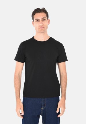 Giovane uomo con capelli corti e scuri, che indossa una semplice t-shirt nera a maniche corte e jeans blu scuro, in piedi davanti a uno sfondo bianco.