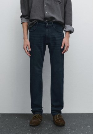 Personne portant un jean bleu foncé, des chaussures marron à lacets, et une chemise grise boutonnée avec les manches retroussées jusqu'aux coudes, debout contre un mur uni.