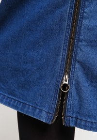 Jupe en denim bleu foncé, dotée d'une fermeture éclair visible sur le côté avec un curseur en métal. Texture lisse et coupe droite.