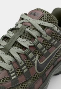 Nike sneaker featuring olive green mesh and leather, pink accents, camo patterns, and flat gray laces with a black Swoosh logo.
