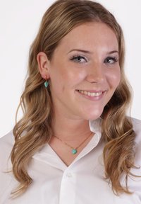 Turquoise stenen oorbellen en een bijpassende ketting bij een vrouw met golvend blond haar, die een witte button-up shirt draagt tegen een effen achtergrond.