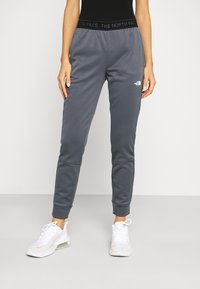 Žena v tmavě šedých teplákových kalhotách The North Face s logem na stehně a elastickým pasem, doplněná o bílé sportovní tenisky.