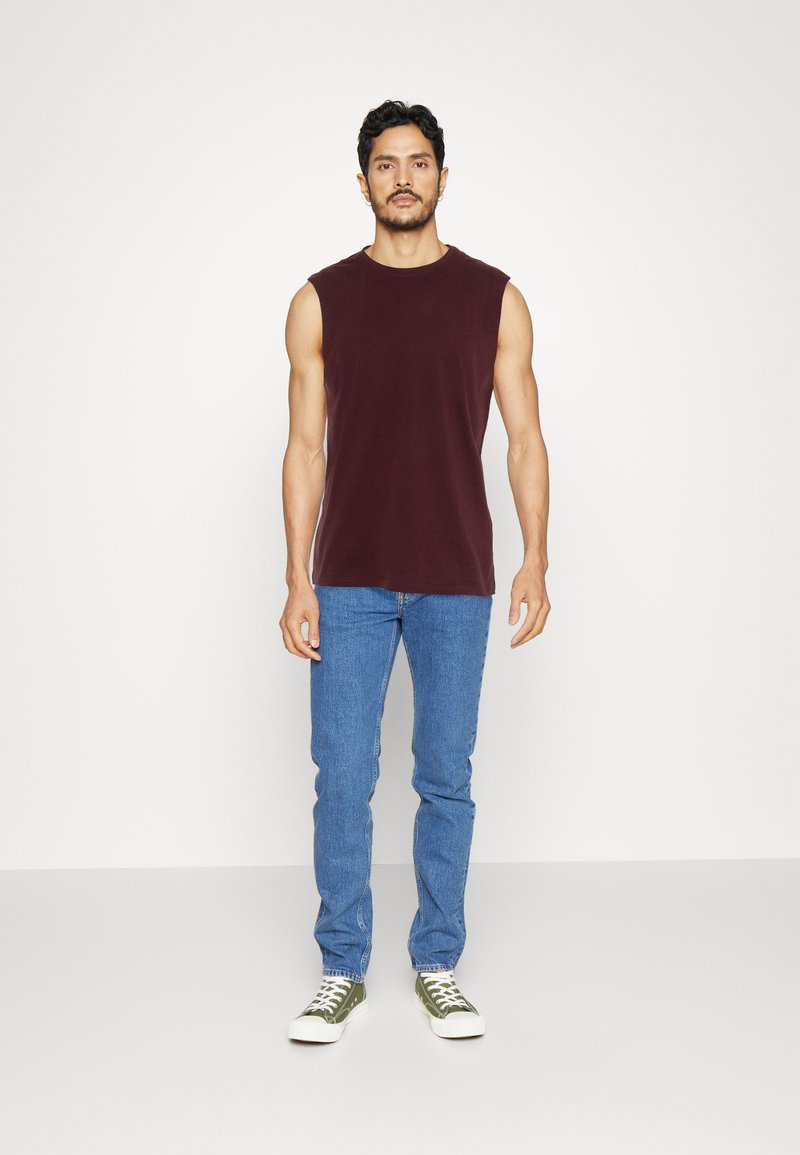 Burgundi ujjatlan póló kék denim farmernadrággal és zöld alacsony szárú tornacipővel párosítva. A modell egy sima fehér háttér előtt áll.