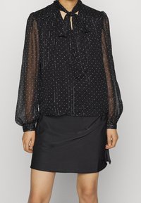 Blusa nera con maniche lunghe trasparenti, motivo a pois e scollo con fiocco. Abbinata a una mini gonna nera in satin.