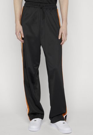 Mustat verkkarihousut oransseilla ja valkoisilla sivuraitoja, joustavalla vyötäröllä ja kiristysnauhalla, päällä valkoiset tennarit.