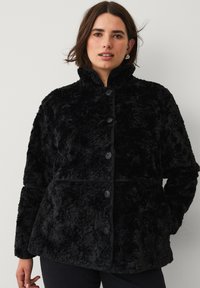 Sort imitation pelsjakke med høj krave, seks frontknapper og en boksformet design. Blød tekstur med vandrette sømme for pasform.