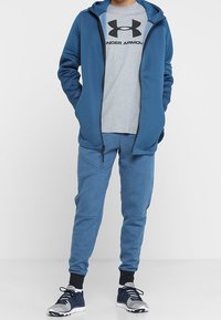 Blå idrottsoverall med en zipparjacka med ribbade detaljer, en grå t-shirt med svart logotyp och matchande blå joggers med mudd.