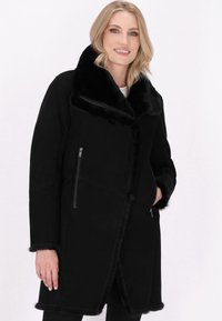 Cappotto in finta pelliccia nera foderato, con ampio colletto, tasche con zip, finitura testurizzata e lunghezza fino al ginocchio.