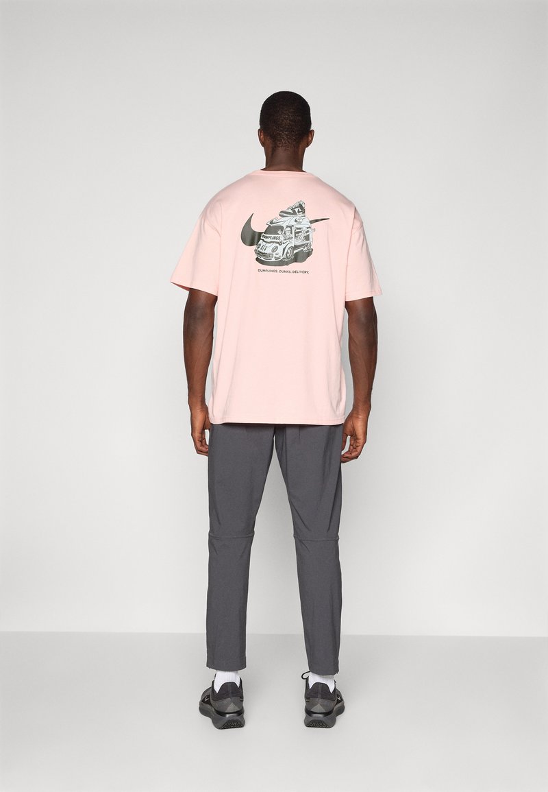 Różowa bawełniana koszulka z grafiką pojazdu dostarczającego jedzenie oraz logo Nike z tyłu. Połączona z szarymi spodniami i czarnymi butami.