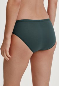 Donkergroene katoenen slips met een gladde textuur, een middelhoog tailleband en elastische afwerking, gezien van achteren met één hand op de dij.
