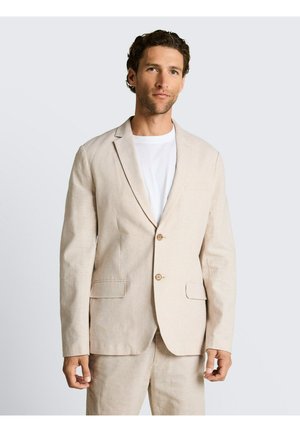 Man die een beige colbert en broek draagt over een wit T-shirt, staand tegen een effen lichtgrijze achtergrond.