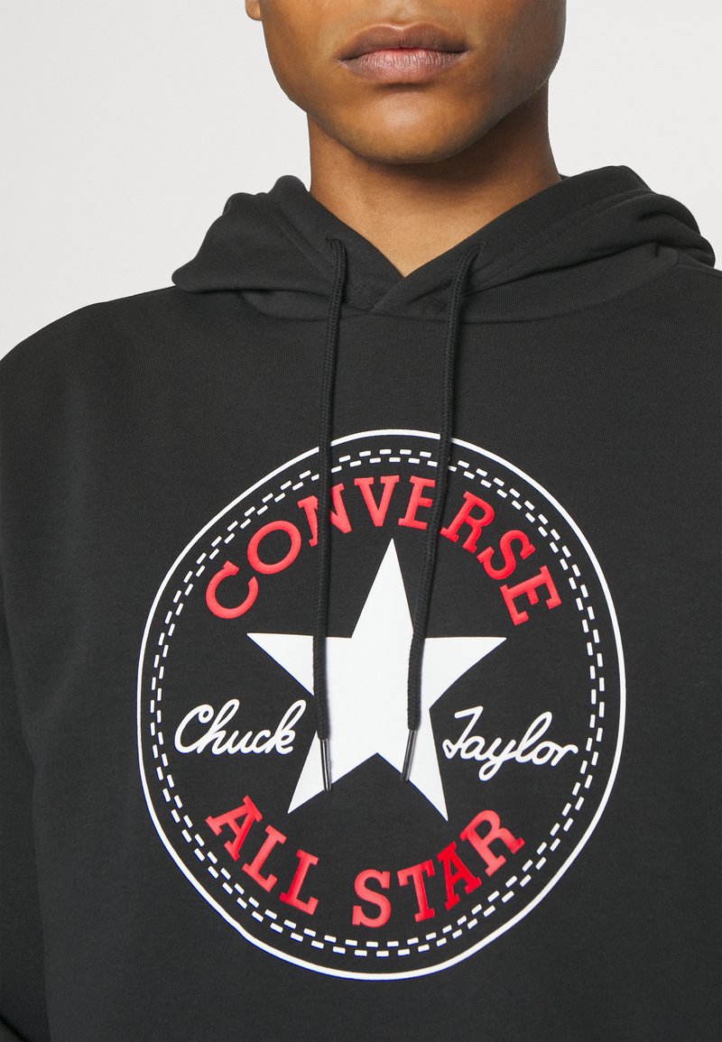 Czarna bluza z kapturem z dużym okrągłym logo z białą gwiazdą. Tekst w czerwieni i bieli brzmi "Converse Chuck Taylor ALL STAR." Teksturowany materiał.