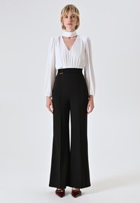 Blusa bianca a maniche lunghe con scollo a V e dettaglio choker, abbinata a pantaloni neri a vita alta e gamba larga. Ballerine burgundy a punta.