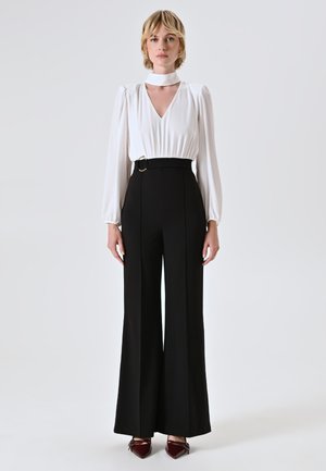Weiße Bluse mit langen Ärmeln, V-Ausschnitt und Choker-Detail, kombiniert mit hochgeschnittenen, weiten schwarzen Hosen. Burgunderfarbene Spitzenflats.