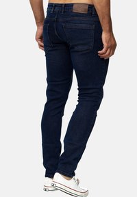 Mørkeblå slim-fit jeans lavet af denim, der har en glat tekstur, baglommer og et mærket lædermærke på taljebåndet. Hvide sneakers synlige.