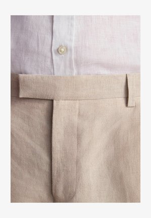 Linen trousers in light beige with a smooth texture and flat front. Complemented by a white linen button-up shirt and a simple waistband.