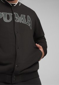 Sweat-shirt noir avec une fermeture à boutons sur le devant, col blanc, et grand logo "PUMA" gris sur la poitrine. Comprend des poignets et une taille côtelés.