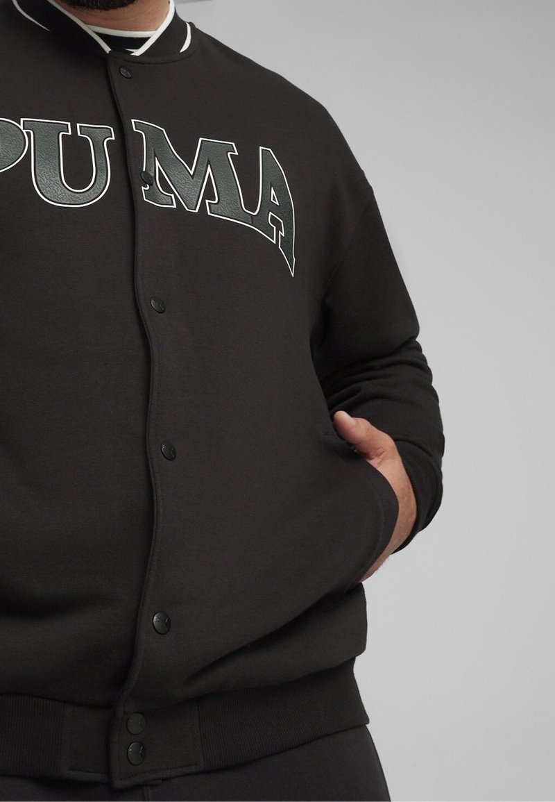 Sweat-shirt noir avec une fermeture à boutons sur le devant, col blanc, et grand logo "PUMA" gris sur la poitrine. Comprend des poignets et une taille côtelés.