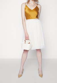 Nainen, joka on pukeutunut sinapinväriseen satiiniseen toppiin, valkoiseen tyllihameeseen ja beigenvärisiin korkokenkiin, pitää pientä valkoista käsilaukkua ja seisoo yksinkertaista valkoista taustaa vasten.