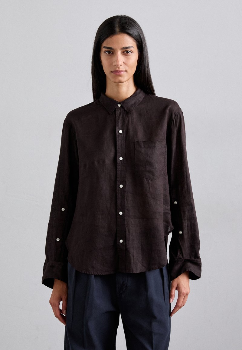 Chemise boutonnée marron foncé à coupe décontractée, col, manches longues, poche poitrine unique et accents de boutons blancs.