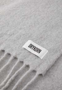 Couverture en laine grise avec des bords à franges, comportant une étiquette en tissu blanc sur laquelle est inscrit "DRYKORN" en lettres noires. Apparence douce et texturée.