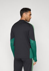 Fekete sportpóló zöld díszítésekkel és hosszú ujjakkal. Texturált oldalsó panelekkel és kontrasztos árnyalatú vállcsíkokkal rendelkezik.