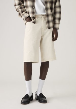 Mann trägt beige kniehohe Shorts, weiße Socken, schwarze Slipper, weißes T-Shirt und braunes kariertes Hemd, die Hände in den Taschen.